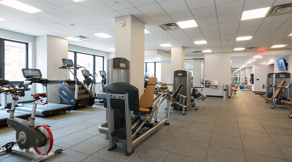 Spacious residential fitness center with grey tile flooring, ceiling lighting, cardio machines, weight equipment, and floor-to-ceiling windows.