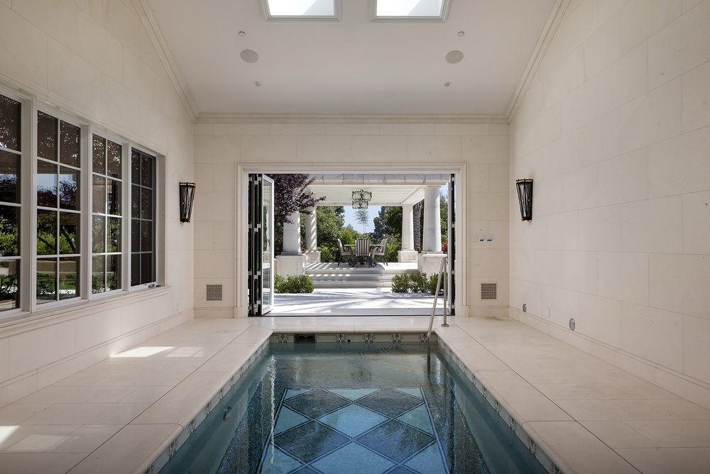 Indoor lap pool room featuring light stone walls, skylights, intricate mosaic tilework, and folding doors leading to a covered patio.