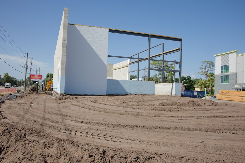 Partially constructed commercial building with concrete block walls and exposed steel frame on a cleared dirt lot, construction equipment nearby suggesting progress, development and urban renovation.