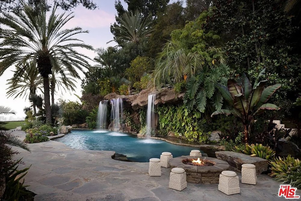 Resort-style backyard swimming pool surrounded by lush tropical landscaping, cascading waterfalls, and a stone fire pit.