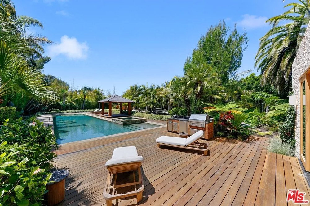 A wooden deck next to a swimming pool features a built-in stainless steel grill, a small sink, and white lounge chairs.