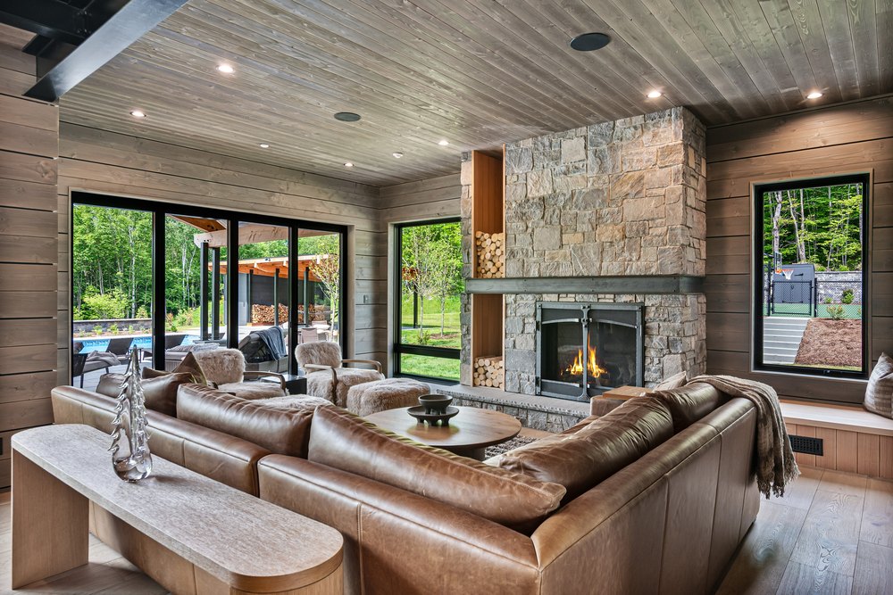 Cozy modern living room featuring a stacked stone fireplace, plush leather sectional sofa, and floor-to-ceiling glass doors.