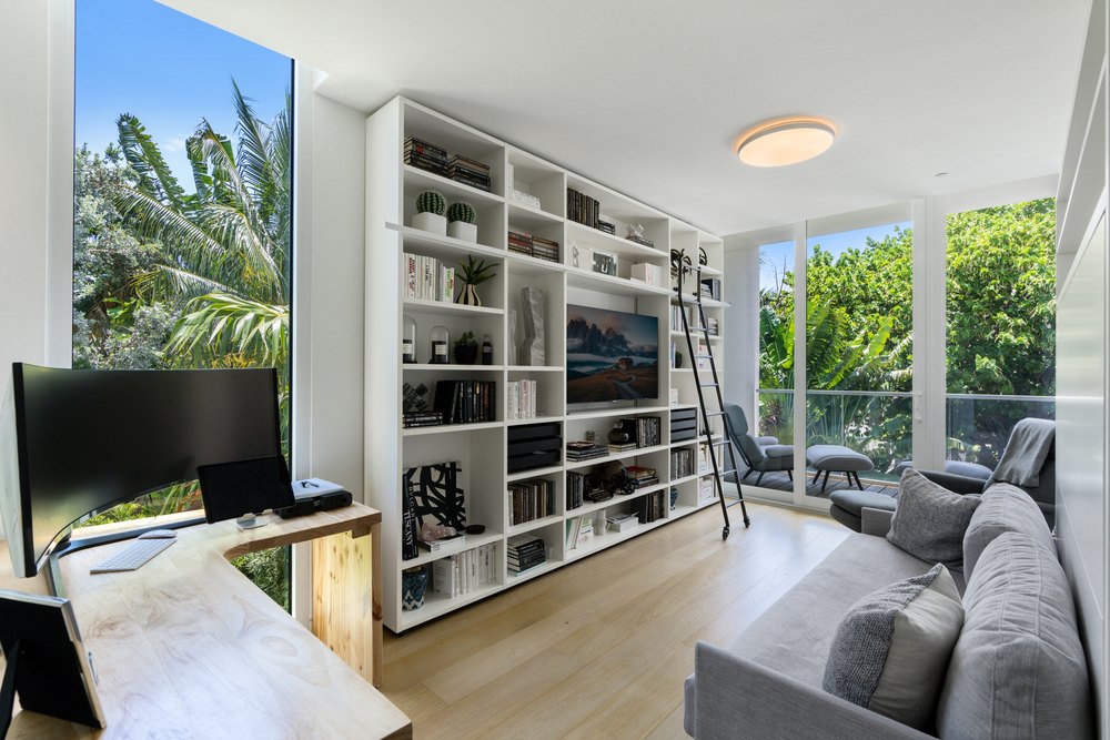 A modern home office with a large white bookshelf and rolling ladder, a wraparound desk, and views of tropical trees.