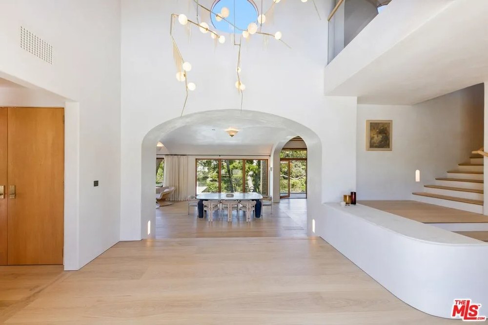 A wide-angle view of a bright entryway shows an arched opening looking into a dining room and a modern branching chandelier.