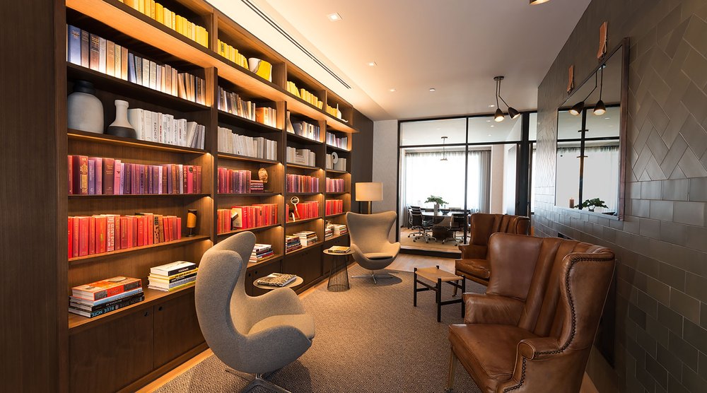 Cozy library reading nook featuring warm integrated bookshelf lighting, grey egg swivel chairs, classic brown leather wingback armchairs, and dark tiled walls.