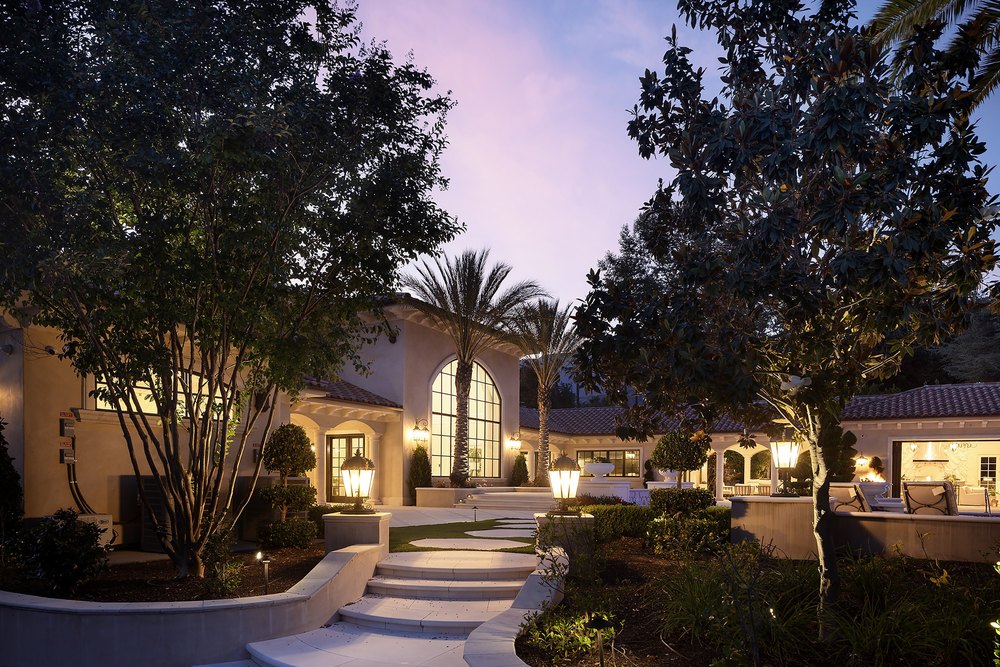 Extensively illuminated stone pathway and garden entry leading toward a Mediterranean mansion under a twilight sky.