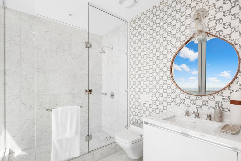 En-suite guest bathroom with marble wall tiles, geometric pattern accent wallpaper, a glass walk-in shower, floating vanity, and a circular wall mirror.