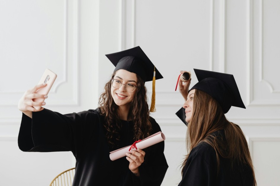 one brunette girl snaps a selfie with her friend, both wearing a black graduation cap, gown, and holding diplomas