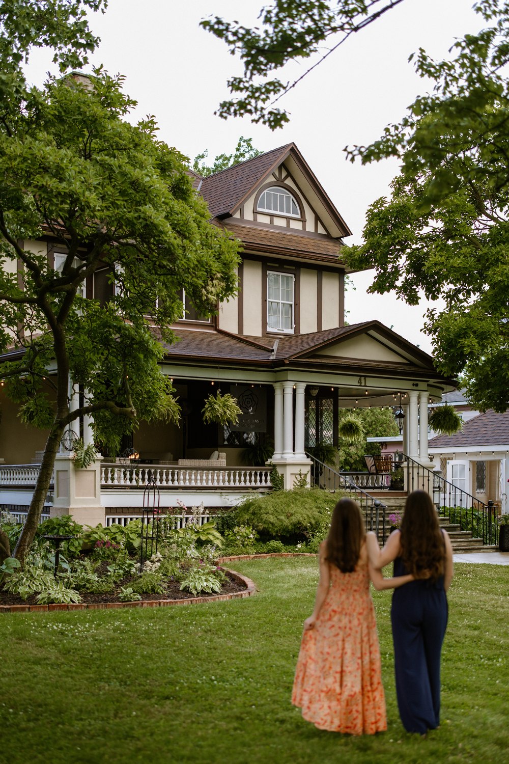 VERTICAL - Kristen and Kathryn Groom walking across a lush green lawn toward The Rose Manor, a grand, historic two-story house with a wrap-around porch.