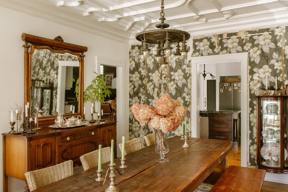 Historic dining room featuring a long wooden table, vintage wallpaper with floral patterns, a large wooden buffet, and an ornate hanging chandelier.