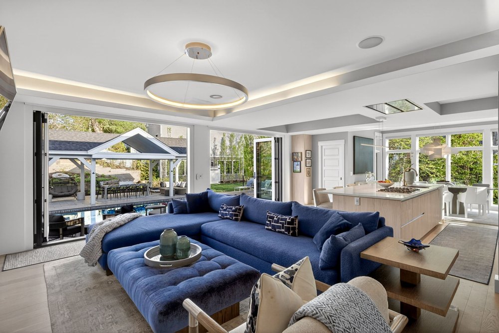 Spacious living room featuring a rich blue sectional, halo pendant lighting, and bi-fold glass doors opening to an outdoor patio and pool.
