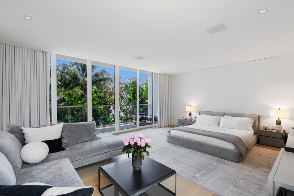 A spacious primary bedroom with a grey bed, a matching sofa, and large windows leading to a balcony.