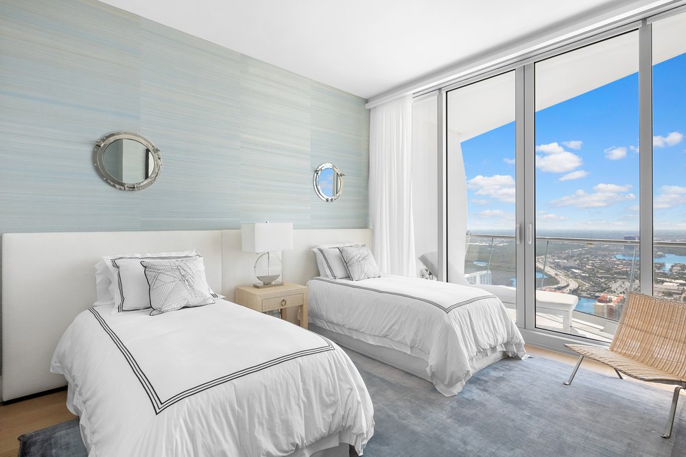 Coastal-inspired twin guest bedroom featuring light blue wallpaper, porthole mirrors, two twin beds with crisp white bedding, and floor-to-ceiling city views.