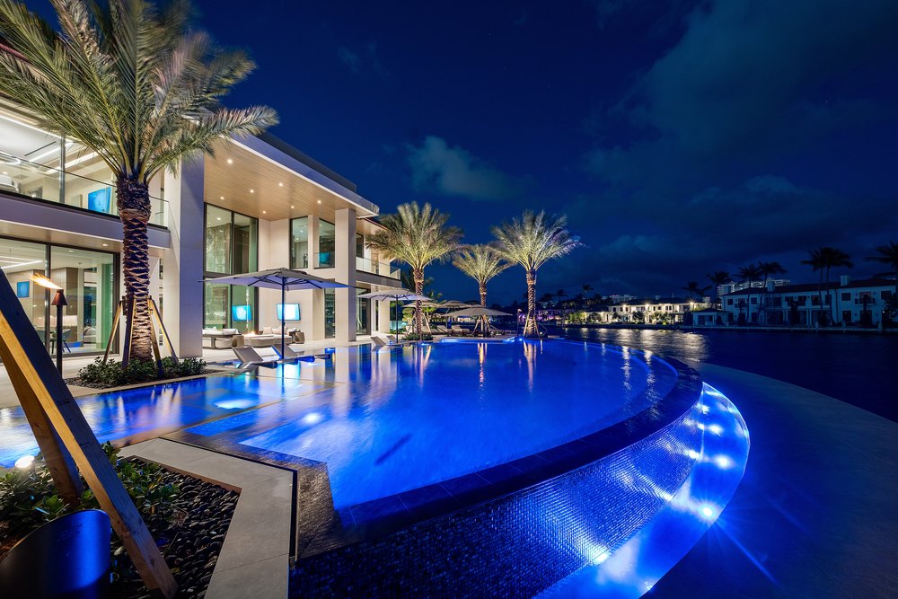Pool Deck Dusk: Blue underwater lighting illuminates a curved infinity pool and modern patio seating next to a contemporary waterfront mansion at night.