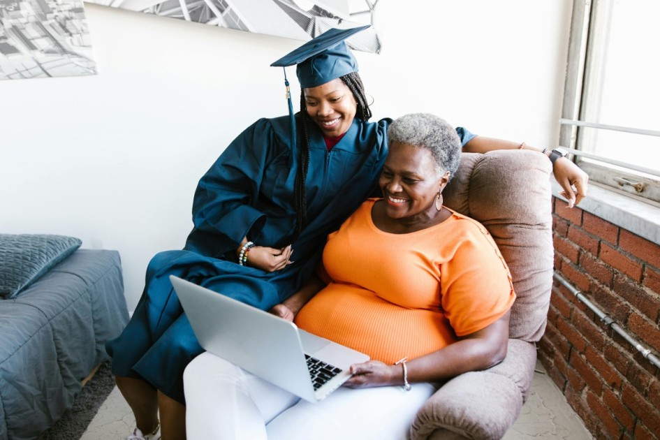 a recent graduate in her cap and gown sits with an older female relative watching a slideshow of her time in school--an idea in Redfin's graduation party ideas article