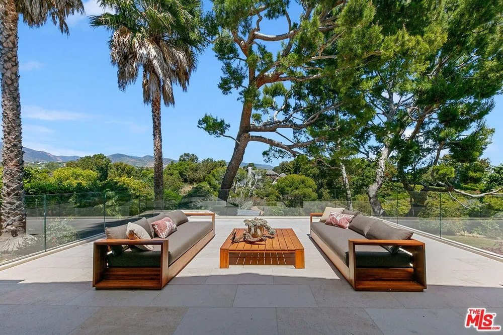 An outdoor stone patio features two large wooden sofas facing each other around a matching coffee table, framed by glass railings and mountain views.