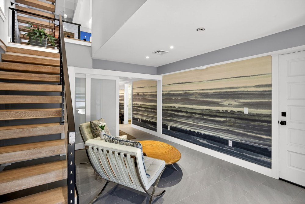 Modern foyer area featuring a floating wooden staircase with cable railings, tiled floors, white Barcelona-style armchairs, and an abstract mural wall.