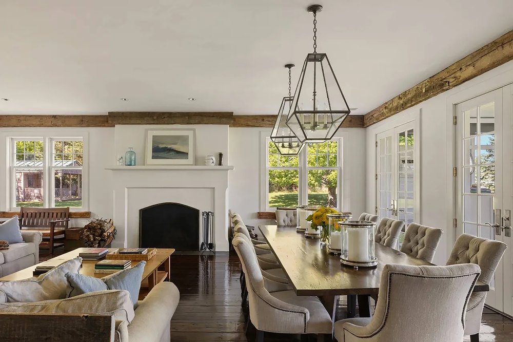 Open-concept living and dining area featuring a wood-burning fireplace, dark floorboards, exposed ceiling beams, a long dining table, and french doors.