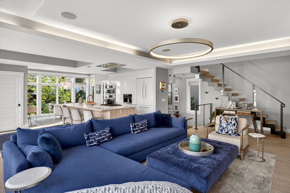 Contemporary open-plan living room with plush blue sectional sofa, tufted ottoman, sleek halo chandelier, and floating wooden stairs.