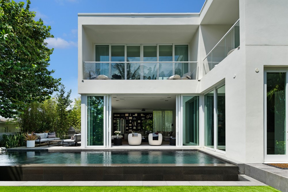 A backyard view showing a dark-tiled lap pool situated right next to the open sliding doors of a modern living room.