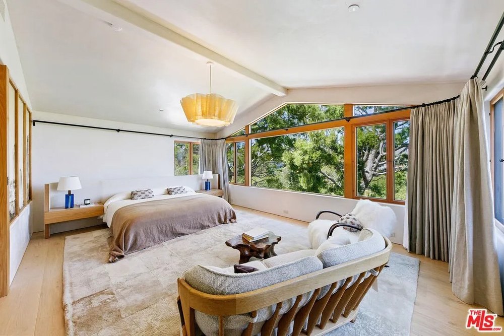 A large bedroom with a vaulted ceiling features a king-sized bed, a plush area rug, and a wide window overlooking a pine forest.