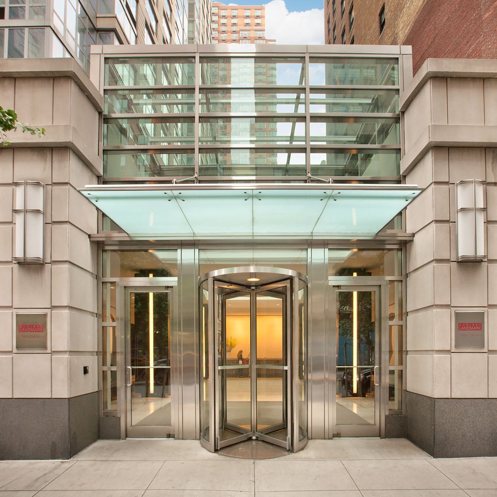 Modern luxury apartment entrance featuring a glass-canopied revolving door framed by stone pillars, sleek metal trim, and contemporary sconce lighting.