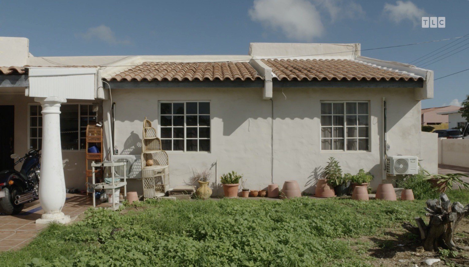 Behind a stucco fence are several wicker plant stands, about a dozen terracotta pots, and a tree stump surrounded by overgrown green weeds.