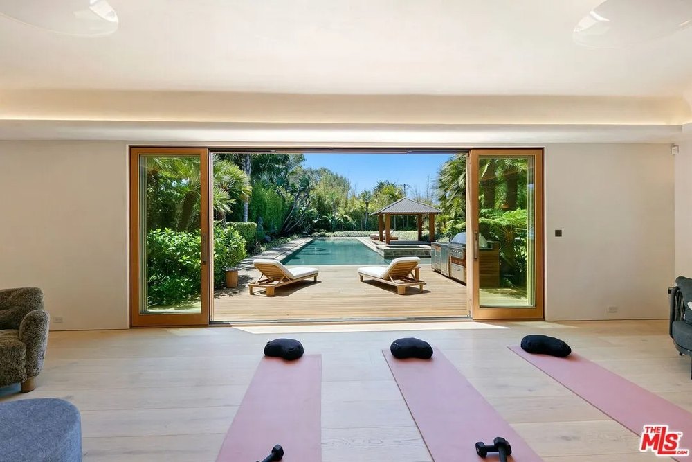 Three pink yoga mats with black cushions are laid out on a light wood floor facing large glass sliding doors that open to the pool deck.