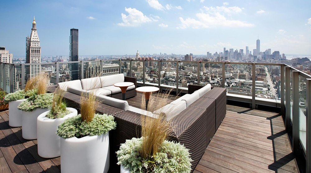 Sunlit rooftop deck featuring dark wicker sectional seating, modern round coffee tables, illuminated planters, and panoramic views of the city skyline.