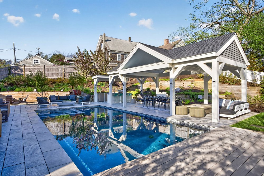 Luxurious backyard oasis with a covered pavilion lounge, outdoor dining set, and an in-ground swimming pool under a bright sky.