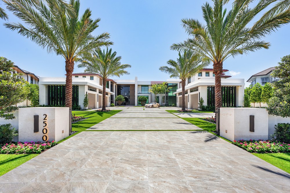 Front Entrance: A wide stone driveway lined with towering palm trees leads to the symmetrical front entrance of the modern white estate.