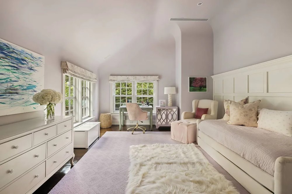 Elegantly appointed bedroom in soft lilac tones featuring a white daybed, plush lavender rug, study desk, and dark hardwood floors.