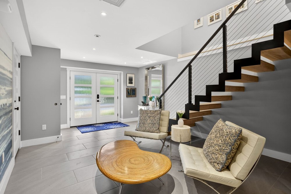 Spacious entry hall with grey tiled floors, double front doors with frosted glass, a kidney-shaped wooden coffee table, and a floating staircase.