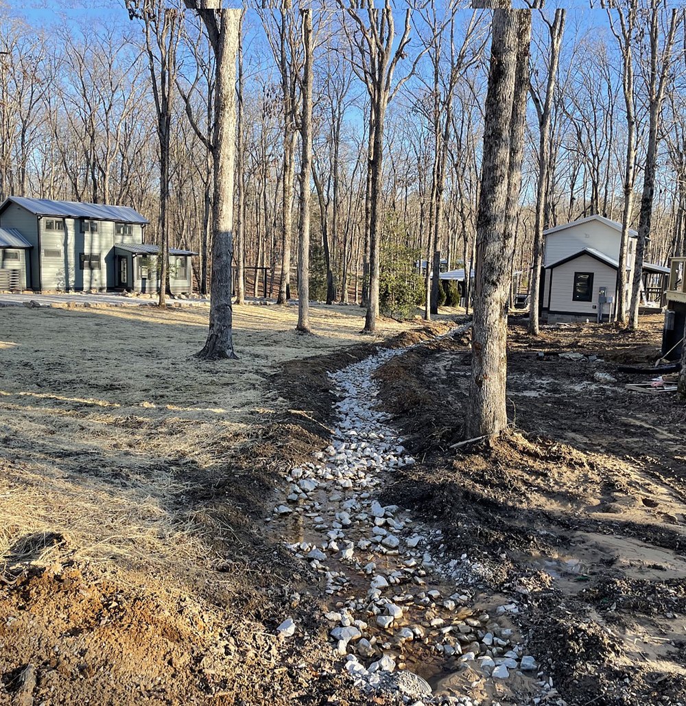 An outdoor residential site under construction featuring a newly installed dry creek bed filled with light-colored riprap stone running between wooded properties and small homes or cabins.