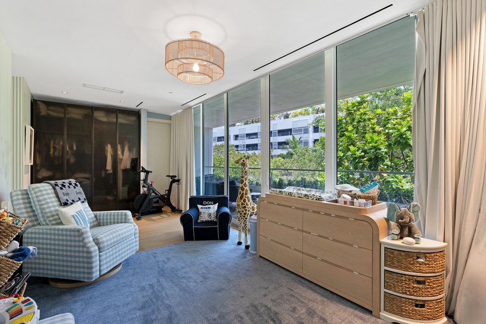A children's bedroom with a wooden dresser, a blue armchair, and large glass walls looking out onto tropical trees.