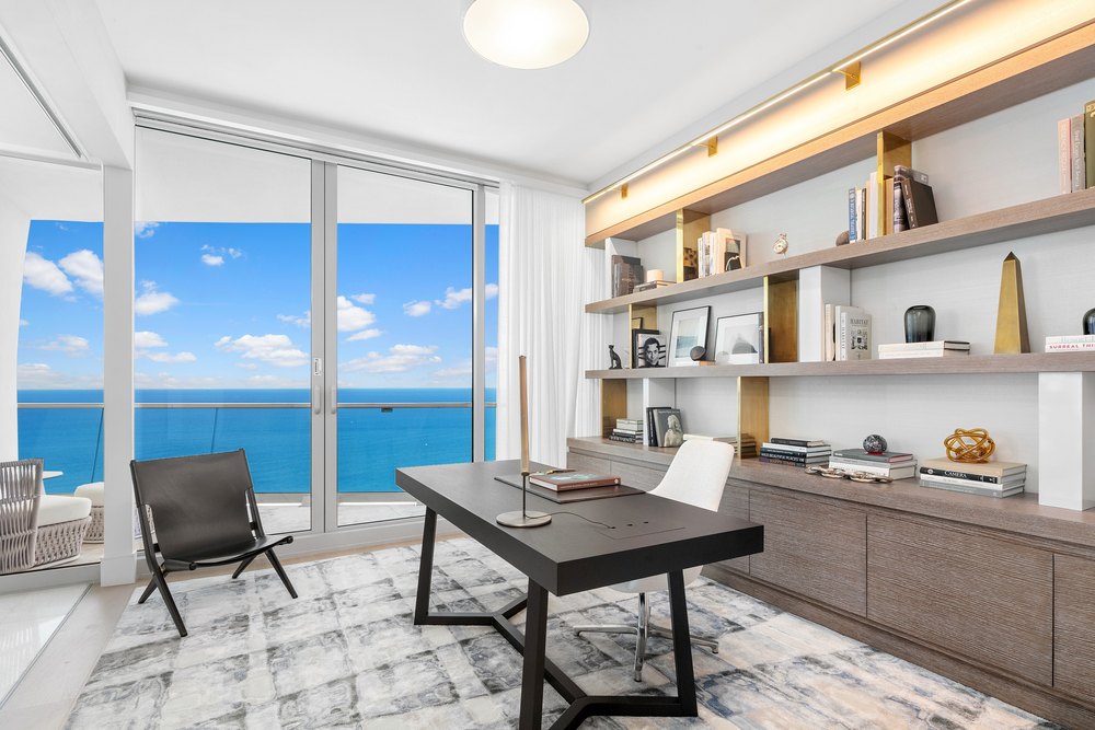 Sleek home office with a dark wood desk, open brass-accented shelving, a modern task light, and floor-to-ceiling sliding glass doors opening to a sea view.