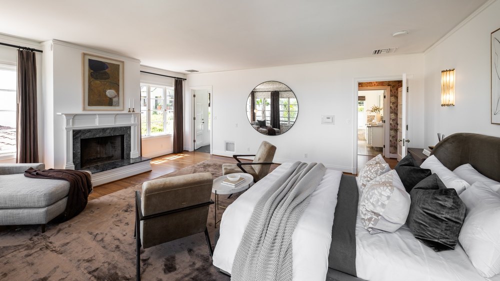 Luxurious primary bedroom featuring a fireplace, a seating area, and a large round wall mirror.