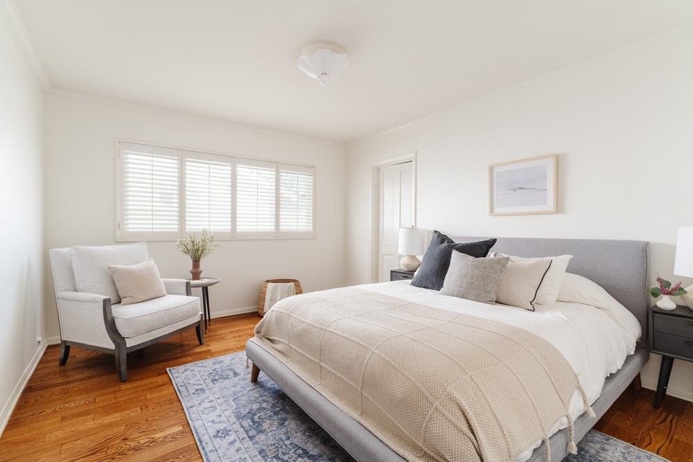 A modern bedroom with a gray fabric bed, a blue patterned area rug, and a white armchair next to a shuttered window.
