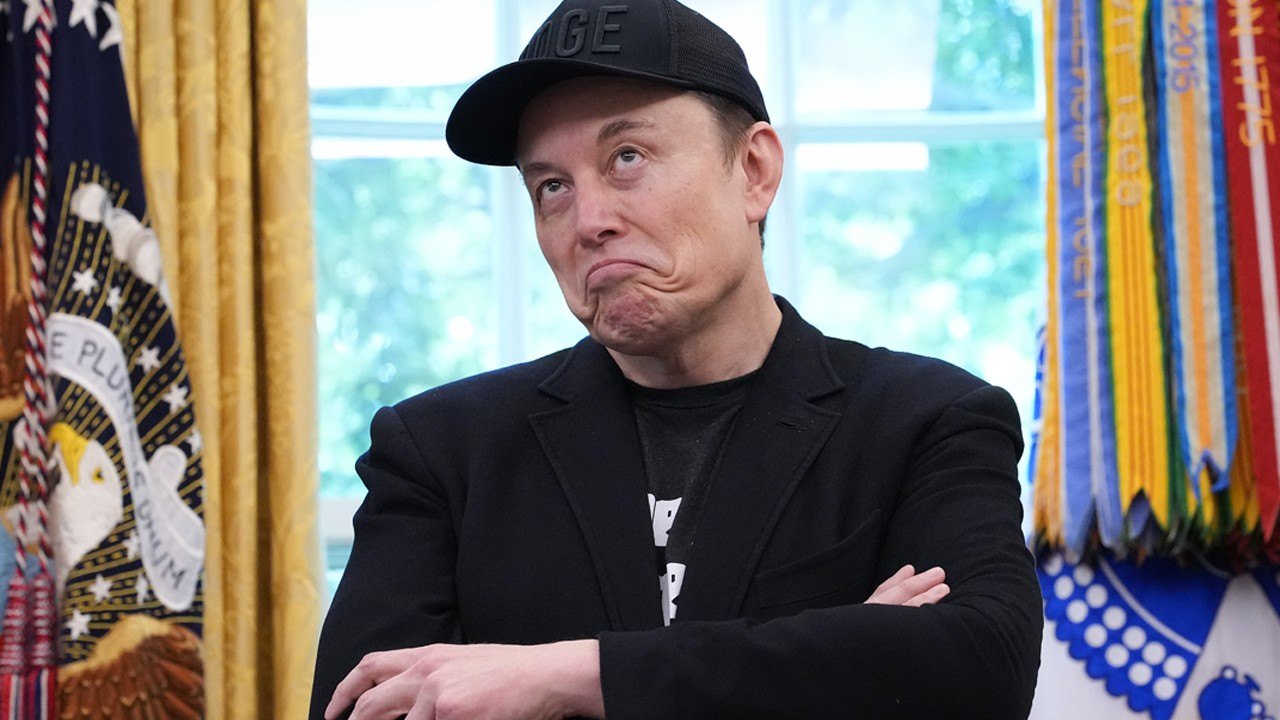 Elon Musk with a black DOGE hat on crossing his arms and looking upward with a frown