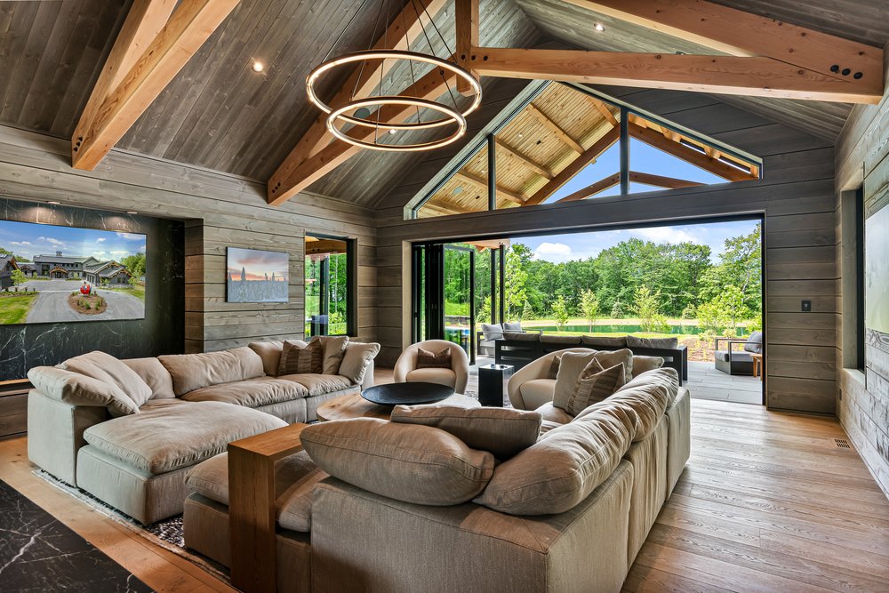Vaulted living room with timber roof trusses, circular ring chandeliers, plush sectional seating, and sliding glass wall doors.