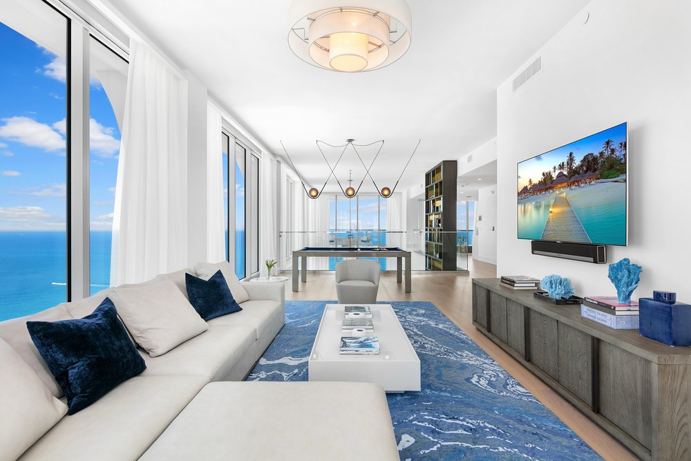Modern penthouse family room with a white sectional sofa, blue accent rug, mounted flat-screen TV, dark wood console, and an open billards space facing the ocean.