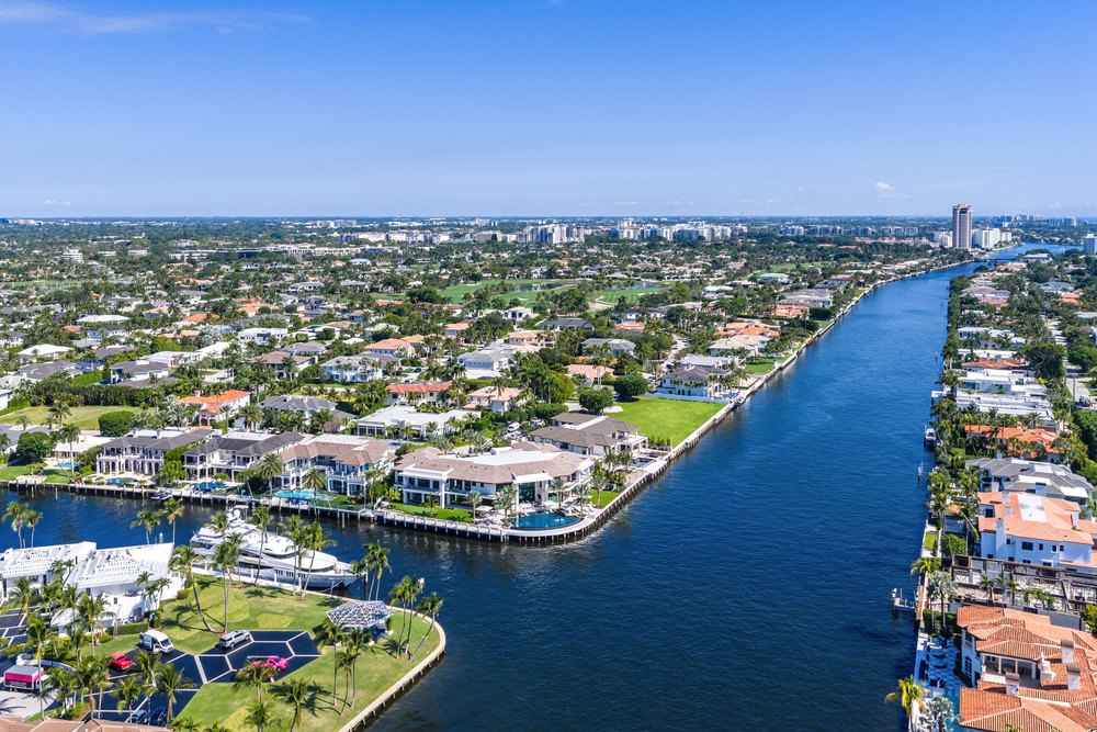 Waterfront Aerial: An aerial perspective shows a luxury waterfront neighborhood with modern estates lining a deep blue coastal canal system under a clear sky.