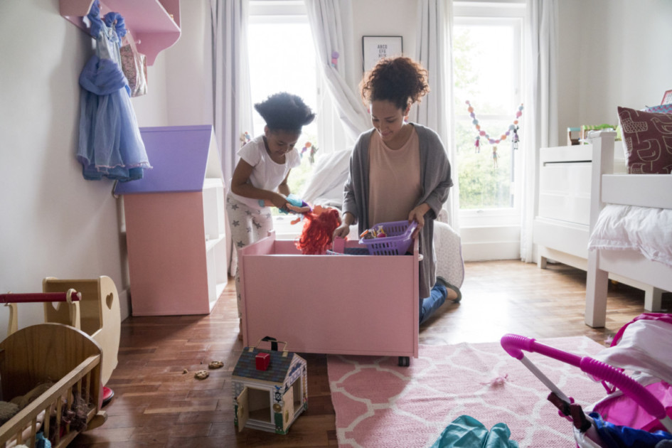 Mother and daughter picking up toys from container. Woman and girl are in bedroom. They are at home.