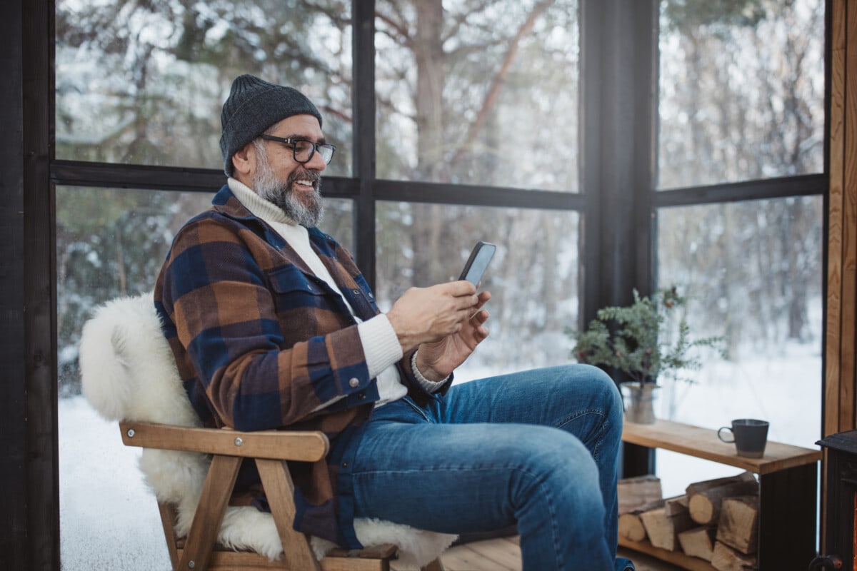 Mature men enjoying in comfortable arranged greenhouse with fireplace. He is using smart phone.