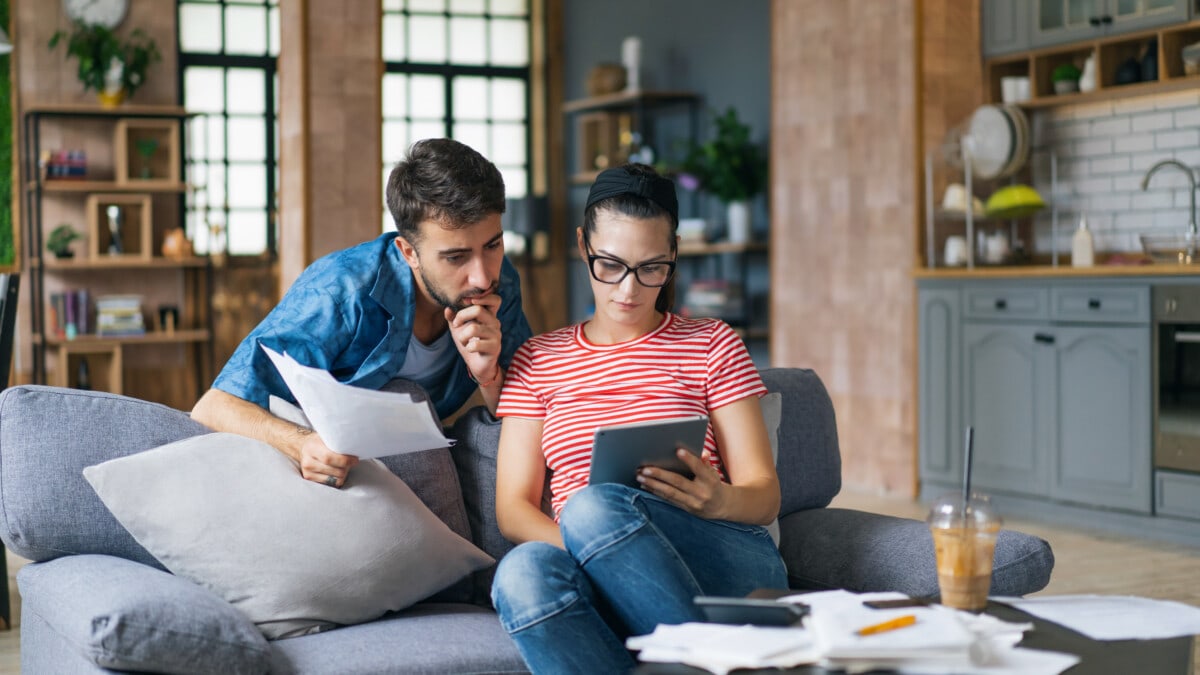 Couple calculating bills at home using laptop and calculator. Young couple working on computer while calculating finances sitting on couch.