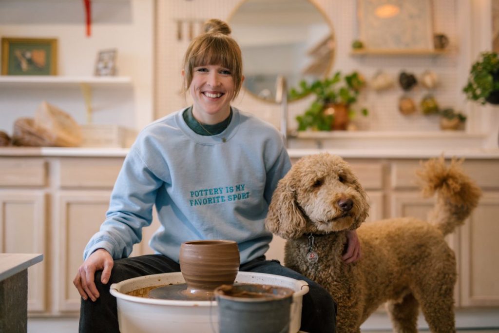 A potter and their large, curly dog sit at a pottery wheel in a gorgeous, light-filled studio.