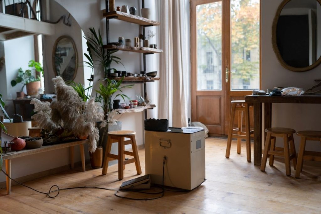 Custom shelving, billowy curtains, house plants, and big mirrors helps this pottery studio feel like home. A pottery wheel with a block of clay ready to be formed sits in a sunny spot in the center of the room.