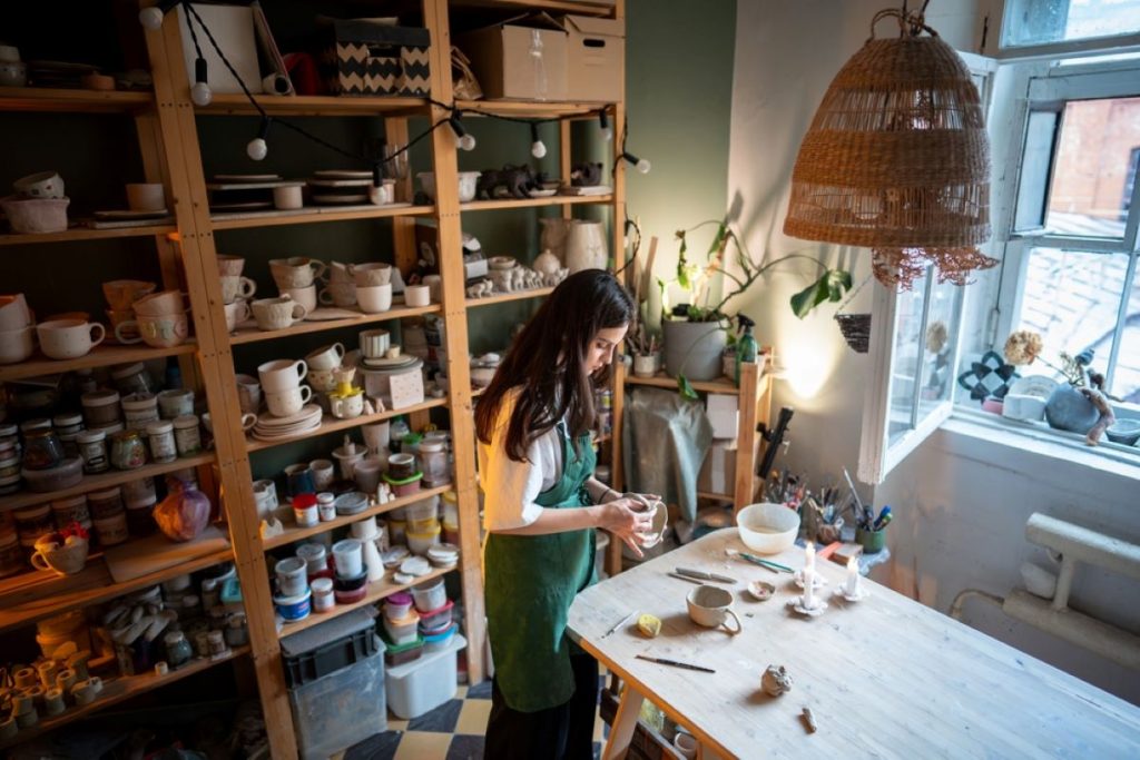 A bohemian-styled home pottery studio that features tall wooden shelves, rattan lighting fixtures, and deep green walls to create a beautiful yet functional space.