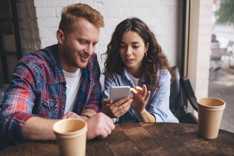House hunting is part of the process when buying a house with a friend. Two friends sit at a coffee shop and look at listings on a phone. 