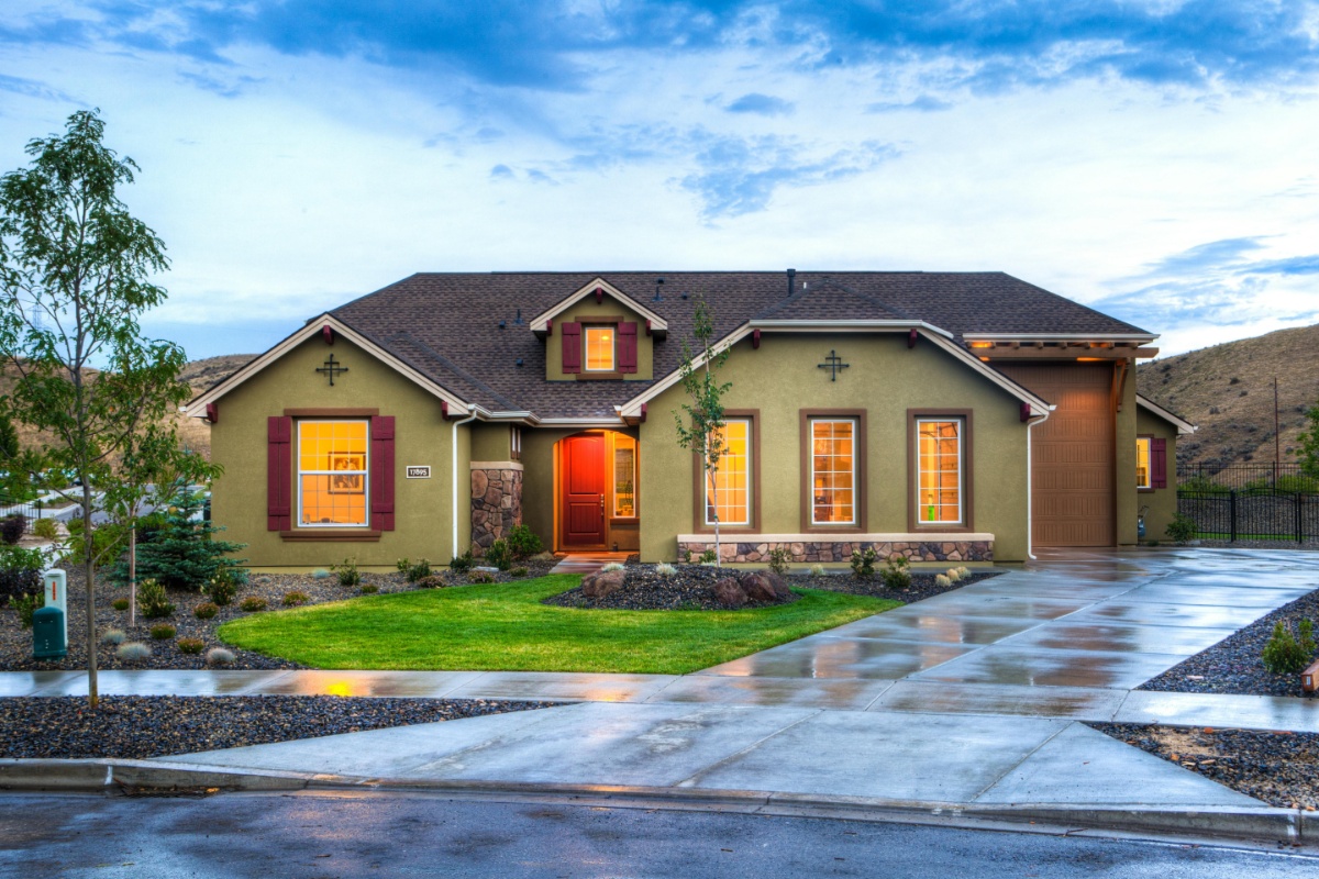 A single-story, olive-green house with a dark brown roof and dark red window shutters stands under a dramatic cloudy sky. The house features multiple windows with warm interior lighting visible, and a bright red front door. A neatly manicured green lawn and a wet concrete driveway lead up to the house. There are young trees and landscaping around the property, and rolling hills are visible in the background under the expansive sky. This image is relevant to the question: Are There Closing Costs if You Pay Cash for a House?"